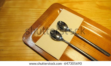 A long silver spoon rests on a wooden tray with tissue paper and all on a wooden table.