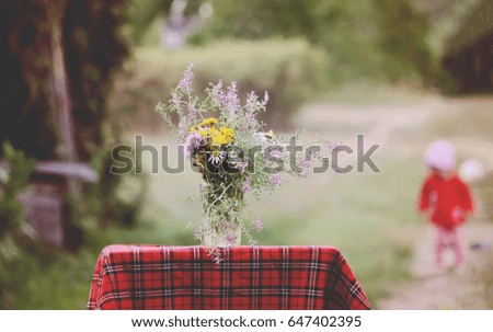 Bouquet of wildflowers on the small table with checkered tablecloth. A little girl playing on the background. Selective focus. Vintage toning.