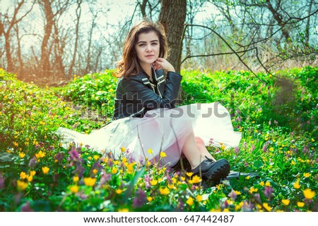 Beautiful teen girl wearing black leather jacket and pink tutu tulle skirt sitting on spring meadow with yellow and purple flowers in the forest 