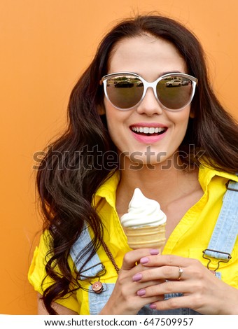 Beautiful  funny young hipster teen girl eating ice cream cone. laughs happy. Bright casual wear, denim shorts. Orange  background, urban style, sunglasses
