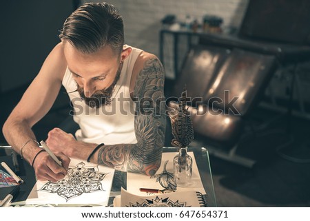 Top view of serene bearded male making tattoo image at desk in apartment. Creativity concept