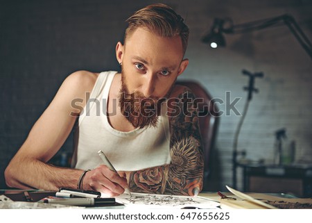 Serene male with tattoo painting image at desk. He looking at camera. Portrait