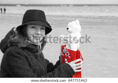A young beautiful girl in a hat sits on the shore of the bay and is happy to receive a teddy bear as a present, a black and white picture with a red color