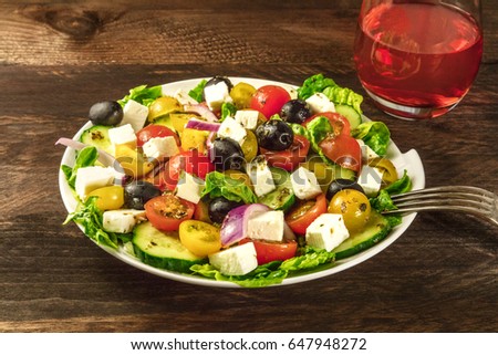 A photo of a plate of Greek salad, with feta cheese, fresh vegetables, and olives, with a glass of rose wine and a fork, on a dark rustic texture with a place for text