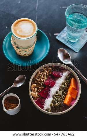 Healthy vegan smoothie bowl made of fresh fruits, pumpkin seeds, nuts, served with coffee latte on black marble table in cafe for breakfast