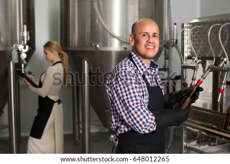 Portrait of smiling mature male brewery worker operating bottling machinery on factory
