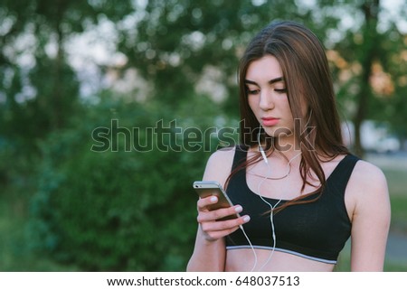 Lifestyle photography outdoors with a girl with an athletic figure who listens to music. Positive athlete with the phone in hand and headphones.