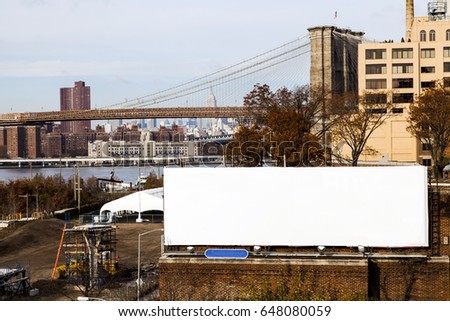 A large blank white billboard located in Brooklyn next to the shore of the East River, with the Brooklyn Bridge and Manhattan seen in the background.