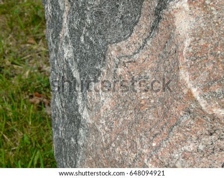 Massive Granite Stone Chunk on Grass in a Park. Natural Red and Dark Grey Abstract Pattern on Untreated Boulder Surface. Architecture, Design, Construction, Geology Concept Texture Background.