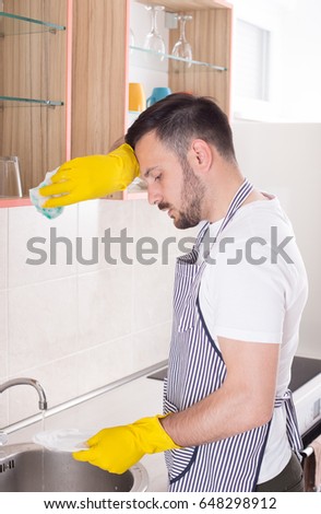 Handsome young man tired of washing dishes and doing chores holding one hand with gloves and sponge on head and clean plate in other