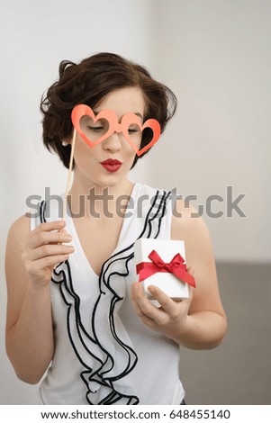 Romantic young woman with a heart shaped glasses party accessory puckering up her lips for a kiss as she looks at a Valentines gift in her hand
