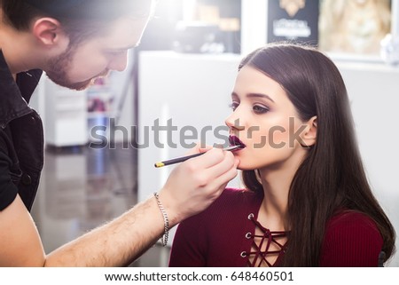 cool male make-up artist Colors the lips brunette girl in a dress in beauty salon