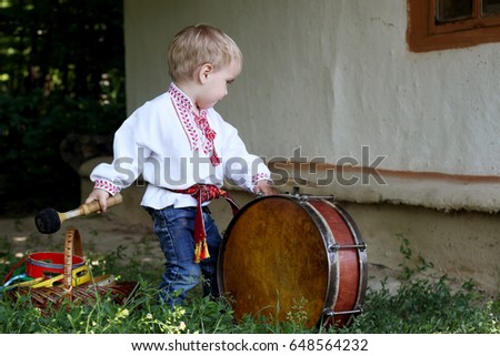 Handsome toddler boy in traditional Ukrainian clothes playing the folk musician instrument boben, Ukrainian history and culture, happy family holiday, summer outdoor portrait