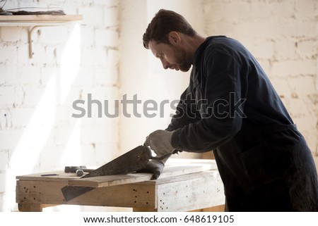 Enthusiast passionate about woodworking hobby cutting wooden board with old-fashioned hand saw on workbench at workshop. Restorer of antique furniture working at new project. Home based business idea