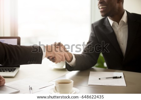 Close up view of handshake between black and white business partners, two smiling multiracial businessmen in formal wear shaking hands over office desk after effective negotiations, signing contract