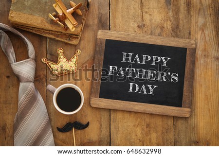 top view image of old books, toy plane, glitter crown, necktie and cup of coffee next to blackboard on wooden background. Father's day concept