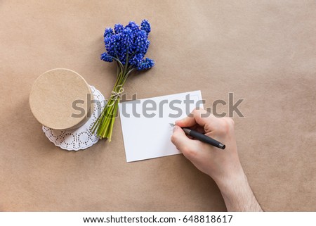 Flatlay blank postcard, man hand writing with the pen, craft present giftbox and the blooming bouquet of muscari (hyacinth) on the craft paper mock up