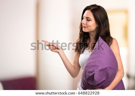 Beautiful young girl with a pillow pointing to the lateral in her apartment