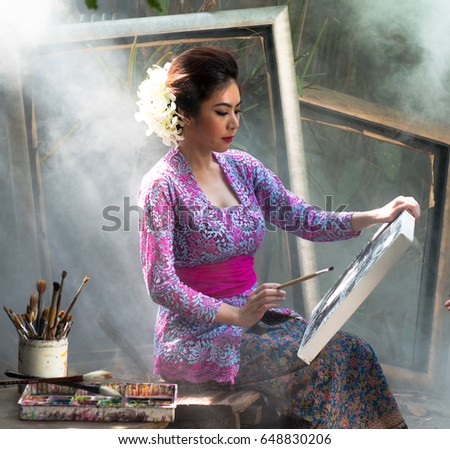 Attactive woman in a vivid color native dress sitting with canvas  in a garden under a beautiful beam,The girl is engaged hold canvas for painting in warm, Young artist painting an autumn landscape