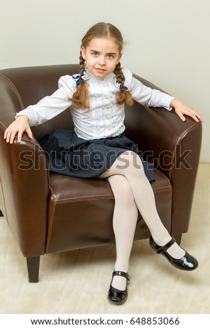 A sweet blonde with long wattled hair in pigtails sits on a brown leather chair and puts her foot on her leg.