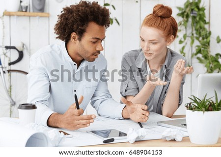 Two architect workers sitting at their cozy workplace surrounded with many papers and modern electronic devices discussing new construction project with emotions using gestures and body language Royalty-Free Stock Photo #648904153