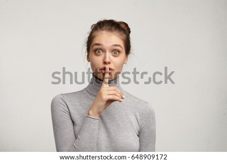Studio shot of wide eyed pretty girl in turtleneck asking for silence or secrecy, holding finger at lips. Cute mysterious young woman saying shh while keeping her pregnancy a secret. Body language