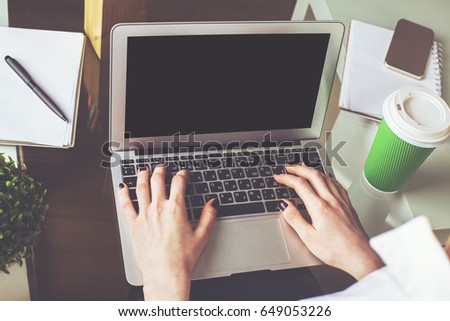 Close up of woman's hands typing/using empty laptop placed on wooden desktop with coffee cup, smartphone and other items. Mock up