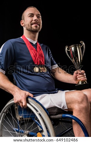 smiling paralympic in wheelchair with gold medals on neck holding champion goblet isolated on black  