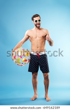 Full length portrait of a happy excited guy holding beach ball and showing thumbs up gesture isolated over blue