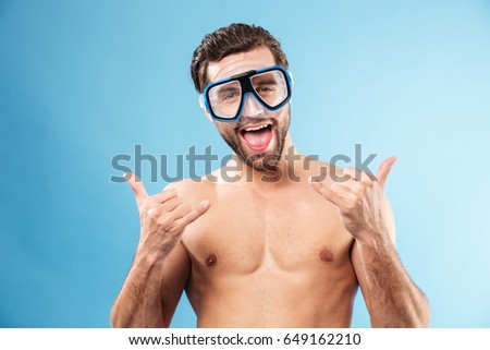 Excited smiling man in swimming goggles showing thumbs up gesture isolated over blue