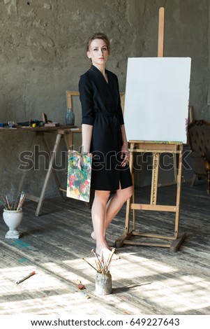 Portrait of elegant woman painter in art studio near empty canvas mockup, holding palette with oil paints and paintbrushes and smiling. Painter at home or workshop. Creative concept