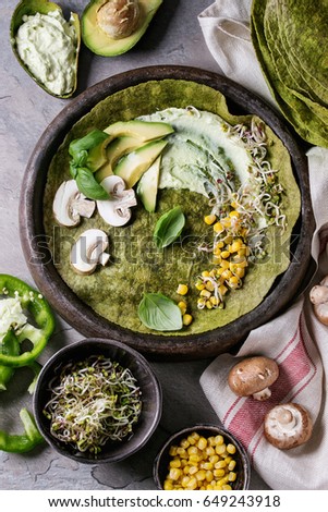 Green spinach matcha tortilla with vegan ingredients for filling. Sweet corn, avocado, green paprika, sprouts, mushrooms served in terracotta plate over gray texture background and textile. Flat lay