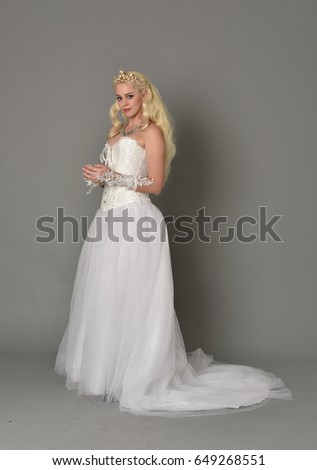 Full length portrait of a blonde woman wearing full length white gown and corset with a crown, standing against a grey studio background.