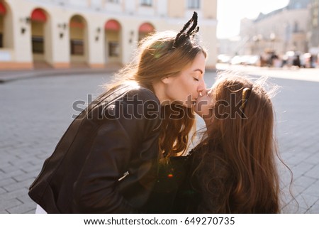 Pretty young woman with eyes closed and happy face expression kissing charming dark-haired daughter. Portrait of fascinating little girl spending time with mother and sitting on the main city square.
