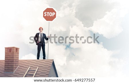 Engineer man standing on house roof and holding red prohibition sign. Mixed media