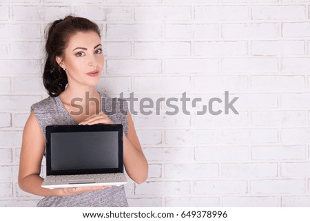 young beautiful woman showing laptop with blank screen over white brick wall