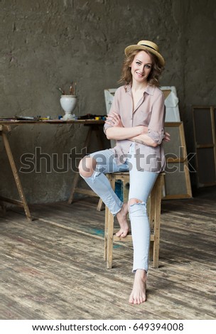 Portrait of a young smiling woman in straw hat sitting on the chair in the artist studio, posing. Easel, frames and paintbrushes on background. Painter workplace interior.