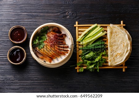 Peking Duck in bamboo steamer served with fresh cucumber, green onions, cilantro and roasted wheaten chinese pancakes with sauce Hoysin on black burned wooden background