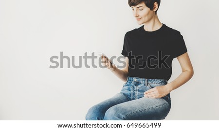 Closeup of young woman wearing blue jeans and using smart phone while sitting on a chair at home, mock-up of black t-shirt, white wall on the background