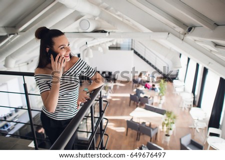 Young woman talking on phone with a friend and smiling while looking away. Blurred cafe, big windows in a background.