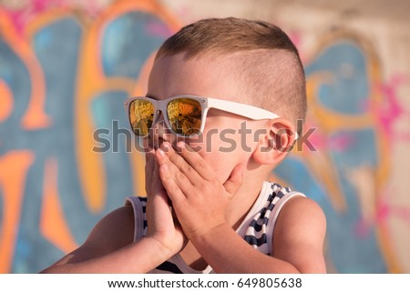 amazed little boy wearing sunglasses and striped shirt closed his mouth with hands looking aside sitting on graffiti background