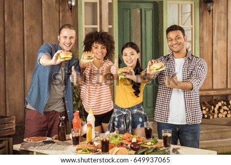 young smiling multiethnic friends holding burgers and showing thumbs up at picnic