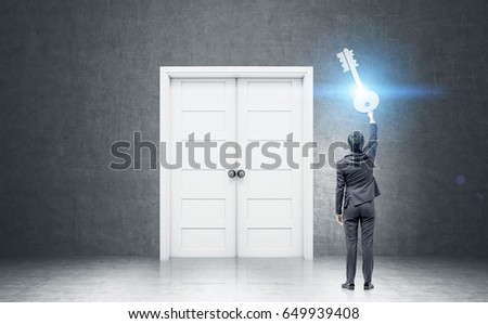 Rear view of a businessman in a dark suit holding a giant key standing near a black wall with a large closed door. Toned image