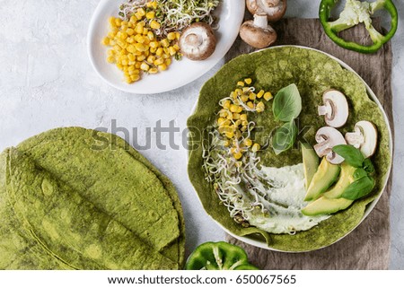Green spinach matcha tortilla with vegan ingredients for filling. Sweet corn, avocado, green paprika, sprouts, mushrooms served in white plate over gray texture background and textile. Flat lay