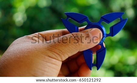 Visible hand held a blue metal Hand spinner. Fidget toy for increased focus, stress relief in an green background with bokeh.