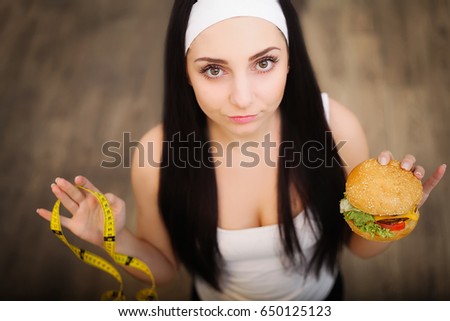 A young woman holding a burger and a measuring tape. A girl stands on a wooden background. The view from the top. The concept of healthy eating.