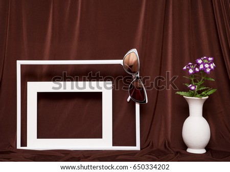 Two empty picture frames, glasses and vase with flowers on background of brown cloth.