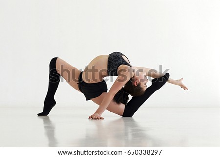 Young slim woman in black sporty outfit doing gymnastics. Girl stretching in bright gym.