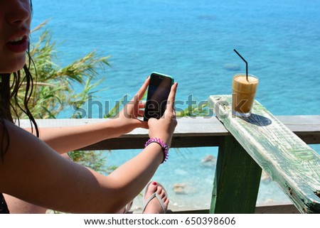Young teenage girl refreshing in cafe with sea view and photographing her ice coffee frappe by mobile phone