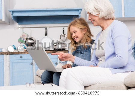 Mature grandmother and her little granddaughter sitting on sofa and looking on screen of laptop computer. Happy girl showing her granny some pictures or photos.
